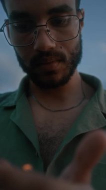 Yanan çakmak ya da mum tutan, rüzgarda titreyen alevi izleyen, gözlüklü genç siyahi adamın yüzünün ve ellerinin dikey yakın çekimi.