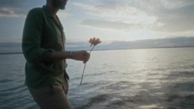 Kumlu göl sahilinde çıplak ayakla yürüyen, parlak turuncu papatya çiçeğini taşıyan ve yalnız hisseden düşünceli genç bir adamın orta ölçekli görüntüsü.