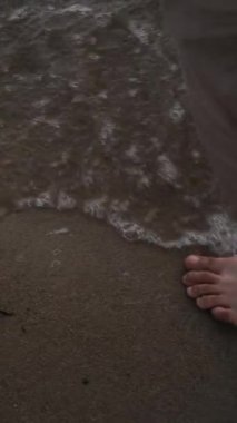 Bej pantolonlu, deniz ya da okyanus kıyısındaki dalgalarda kumda yalın ayak yürüyen, teninde serin su hisseden anonim bir adamın bacaklarının dikey görüntüsü.