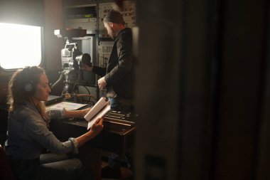 Kulaklıklı genç bir kadın kağıt belgelerle mesajlara bakarken aynı zamanda ses karıştırıcı konsolda meslektaşlarına karşı oturuyor.