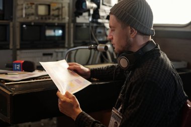 Ciddi bir erkek operatör, TV minibüsündeki ses karıştırma konsolunda otururken çalışan kağıt dökümanı üzerinde çalışıyor