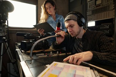 Genç erkek operatör, kadın meslektaşına iş yerinde yardım ederken konsoldaki sesi ayarlıyor ve mikrofonla konuşuyor.