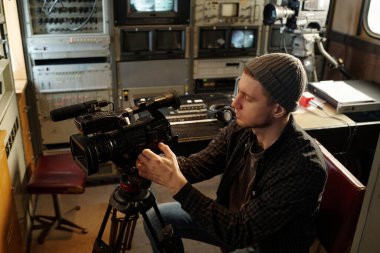 Genç adam iş yerinde mikrofon ve ses karıştırıcı konsolla otururken profesyonel video kameranın odak noktasını ayarlıyor