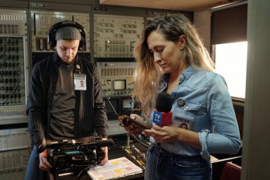 Mikrofonlu genç muhabir iş yerinde dururken akıllı telefon kullanıyor ve erkek operatör çekim için hazırlanıyor.