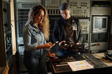 Genç bayan gazeteci cep telefonuyla mesajlaşırken erkek operatörün yanında kamerayı ayarlıyor.