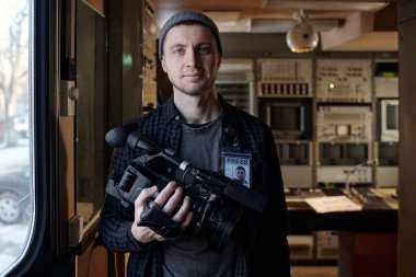 Elinde profesyonel kamera olan genç ve başarılı bir erkek operatör TV minibüsünün içinde dururken sana bakıyor.