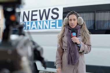 Haber kanalının TV minibüsünün yanında duran ve konuşma sırasında video kameraya bakan kendine güvenen genç kadın muhabire odaklan.