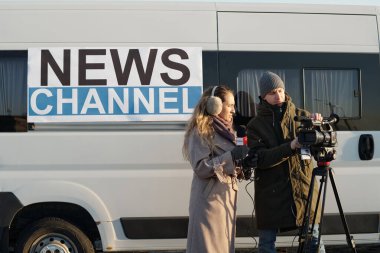 Erkek operatörün yanında duran genç bayan gazeteci haber çekmeden önce profesyonel video kamerayı ayarlıyor.