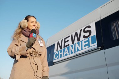 Genç, gülümseyen bir kadın mikrofonla konuşuyor. Açık hava haber çekimi sırasında son gelişmeleri aktarıyor.