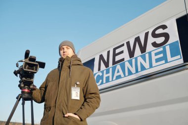 Gündelik kıyafetli genç adam TV minibüsüne karşı profesyonel video ekipmanının yanında dururken kameraya bakıyor.