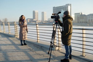 Genç adam, mevcut durumu açıklayan kadın gazeteciyi çekerken tripodda profesyonel video kameranın önünde duruyor.