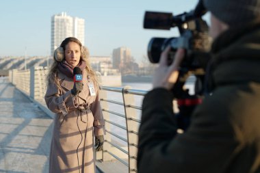 Sıcak bej ceketli ve kulaklıklı genç bir kadın mikrofonla konuşurken son gelişmeleri bildiriyor.