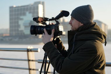 Yeni çekimlere hazırlanırken, gülümseyen ceketli ve bereli genç adamın video kamerayı ayarlarken yan görüntüsü