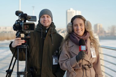 Günlük giysiler içinde genç bir erkek ve kadın gazeteci televizyon kanalı için haber bülteni çekerken sizi izliyor.