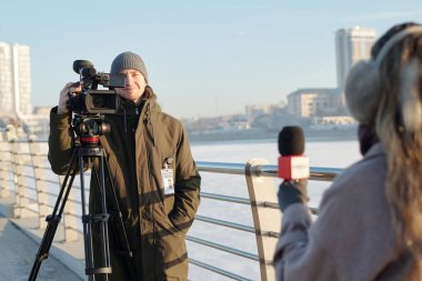 Mutlu genç adam elinde mikrofonla gazetecinin önünde dikilirken profesyonel video kamerayla haber sunuyor.