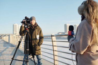 Nehir kenarında durup kadın gazeteciyi çekerken profesyonel video kamera kullanan sıradan giyinmiş genç bir adam.