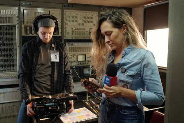 Mikrofonlu genç muhabir iş yerinde dururken akıllı telefon kullanıyor ve erkek operatör çekim için hazırlanıyor.