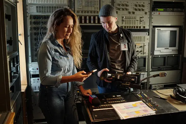 Genç bayan gazeteci cep telefonuyla mesajlaşırken erkek operatörün yanında kamerayı ayarlıyor.