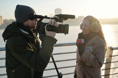 Profesyonel video kameralı genç adam mikrofonlu kadın gazetecinin önünde dururken yeni bir haber çekiyor.