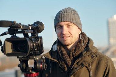 Şehir ortamında video ekipmanlarının başında kameraya bakan gri bereli mutlu genç adam.