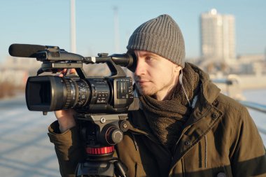 Bereli ve ceketli genç ciddi bir erkek operatör şehir ortamında nehir kenarındaki profesyonel kamerada çekim yapıyor.