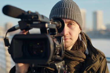 Genç erkek operatör dışarıda röportaj yaparken kameraya bakıyor ve elinde video ekipmanı tutuyor.