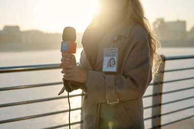 Kamera önünde şehir ortamında nehir kenarına karşı duran mikrofonlu kadın muhabirin görüntüsü.