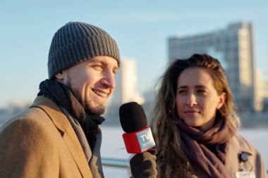 Bereli ve ceketli genç adam kadın muhabirin yanında duruyor ve açık hava röportajı sırasında mikrofonla konuşuyor.