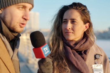 Mikrofonlu genç bir kadın, erkek politikacıya bakıyor. Şehir ortamında röportaj sorularını yanıtlıyor.