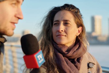 Uzun sarı saçlı genç bayan gazeteci TV kanalının röportajı sırasında mikrofonla konuşan adama bakıyor.