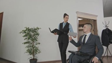 Yavaş çekimde kadın kişisel asistanın ofiste tekerlekli sandalyeye binen erkek yöneticiye dokümanları verirken görüntüsü var.