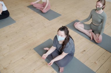 Bir grup maskeli ve aktif giysili öğrenci minderlerde yoga pozisyonunda rahatlama alıştırması yapıyor.