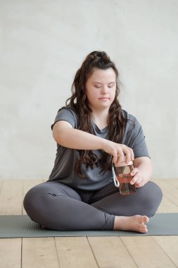 Genç engelli kadın paspasın üzerinde otururken plastik bardağı suyla açıyor.