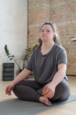 Aktif kıyafetli genç bir kadın antrenman sırasında paspasın üzerinde otururken sana bakıyor.