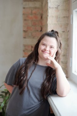 Gri spor giysili mutlu kız pencere eşiğine yaslanırken sana bakıyor.