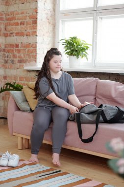 Spor kıyafetli kız kanepede otururken yoga eğitimi için her şeyi büyük bir çantaya koyuyor.