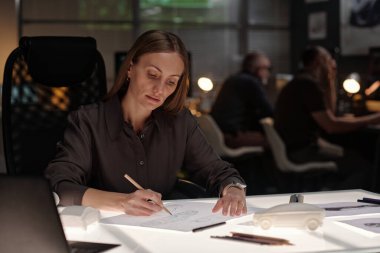 Kalkınma bürosunun kadın mühendisi işyerinde oturuyor ve kağıt üzerinde kalem ile yeni araba modellerinin taslaklarını çiziyor.