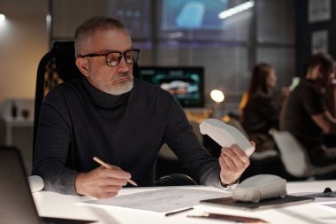 Gözlüklü ve kazaklı kıdemli mühendis büronun yanında oturuyor ve kağıt üzerinde çizerken araba modeline bakıyor.