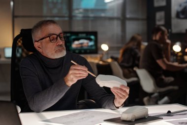 Kendine güvenen olgun erkek otomobil tasarımcısı elinde beyaz araba modelini çizimlerle masasında otururken gösteriyor.