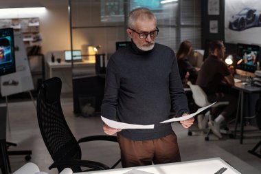 Ciddi deneyimli erkek mühendis en yenilikçi araba modelinin taslağını seçerken işyerindeki kağıtlara bakıyor.