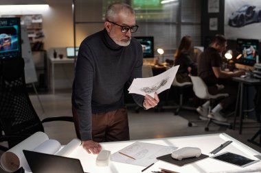 Ciddi yetişkin erkek mühendis, iş sırasında iş yerinde eğildiği sırada yeni araba çizimleriyle kağıtlara bakıyor.