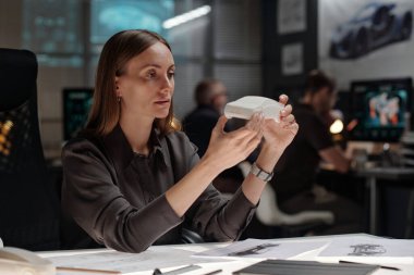 Genç mühendis, beyaz araba modelini tutarken ve incelerken otomotiv tasarım stüdyosunda çizimlerle işyerinde oturuyor.