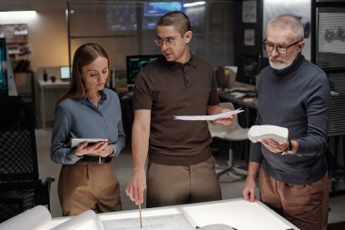 Genç kendine güvenen erkek araba tasarımcısı masadaki kağıt belgeye işaret ediyor ve tartışma sırasında tabletli kadın meslektaşına bakıyor.