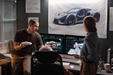 Araba mankenli genç mühendis ve iş yerinde tabletli kadın bilgisayar ekranlarında otomobilleri tartışıyor.