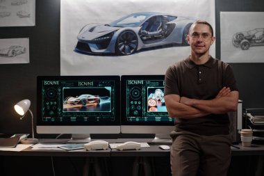 Mutlu genç erkek araba tasarımcısı kollarını göğsüne dolayıp ofisteki bilgisayarların başında dururken sana bakıyor.