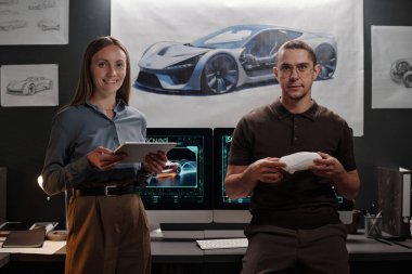 Genç erkek ve kadın araba tasarımcıları iş yerinde bilgisayarlı posterli ve arabalı kameralara bakarken kameraya bakıyorlar.