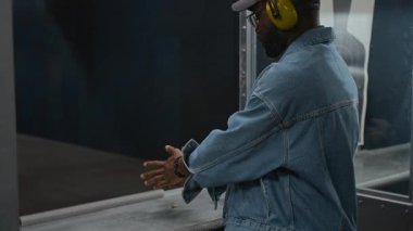 Kot ceketli, kamyoncu şapkalı ve kulaklıklı orta boy Afro-Amerikan adam atış poligonunda duruyor, kurşunları tabanca şarjörüne dolduruyor, tek başına pratik yaparken hedefe nişan alıyor.