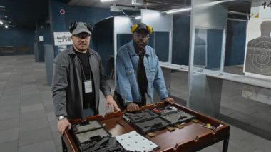 Kafkas atış eğitmeninin orta boy portresi ve Afrika kökenli Amerikalı erkek müşteri masa başında silah, cephane ve mermilerle birlikte kameraya bakıyor.