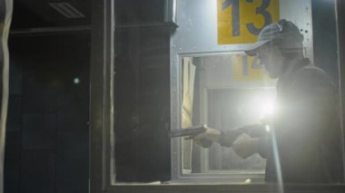 Arka ışıklandırılmış orta ölçekli, kulak koruyucu takmış, yarı otomatik silah yükleyen, hedefe nişan alan ve ateş eden mercek mercekli erkek müşteri, atış sahasının camından ateş ederken kameraya alındı.