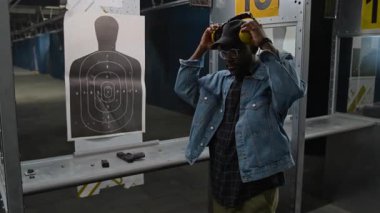 Kafasında kulaklık olan, siluet hedefinin yanında duran ve kameraya bakarken yarı otomatik tabanca kullanan, kendinden emin, Afro-Amerikan poligon müşterisinin orta boy portresi.